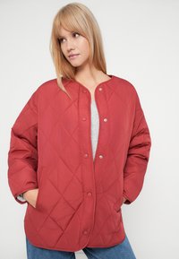 Kvinne med blondt hår iført en knappet, quiltet rød jakke, grå topp og blå jeans med hendene i jakkelommene.