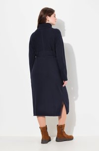 Tummansininen pitkähihaisten neulemekko, jossa on korkea kaulus, teksturoitu takapaneeli, vyötärökiristys ja sivuhalkiot; yhdistettynä ruskeisiin vetoketjullisiin nilkkasaappaisiin.