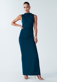 Groene, mouwloze maxi-jurk, aansluitend model, nep col, gladde textuur, split tot halverwege de dij, gecombineerd met naakte sandalen met hak.