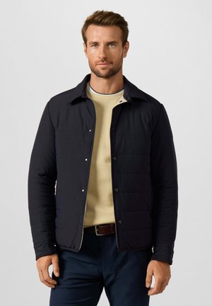 Hombre con una chaqueta negra abotonada sobre un suéter beige y pantalones oscuros, de pie frente a un fondo claro y liso.