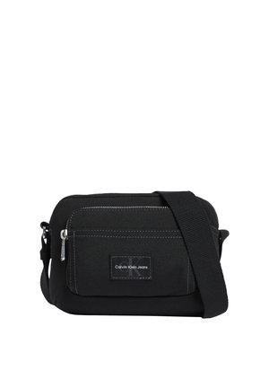 Bolso bandolera de tela negra con bolsillo delantero con cremallera, correa ajustable y parche con el logo de Calvin Klein Jeans en el centro del bolsillo.