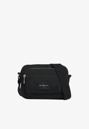 Sac bandoulière en tissu noir avec poche avant zippée, sangle réglable et écusson du logo Calvin Klein Jeans au centre de la poche.