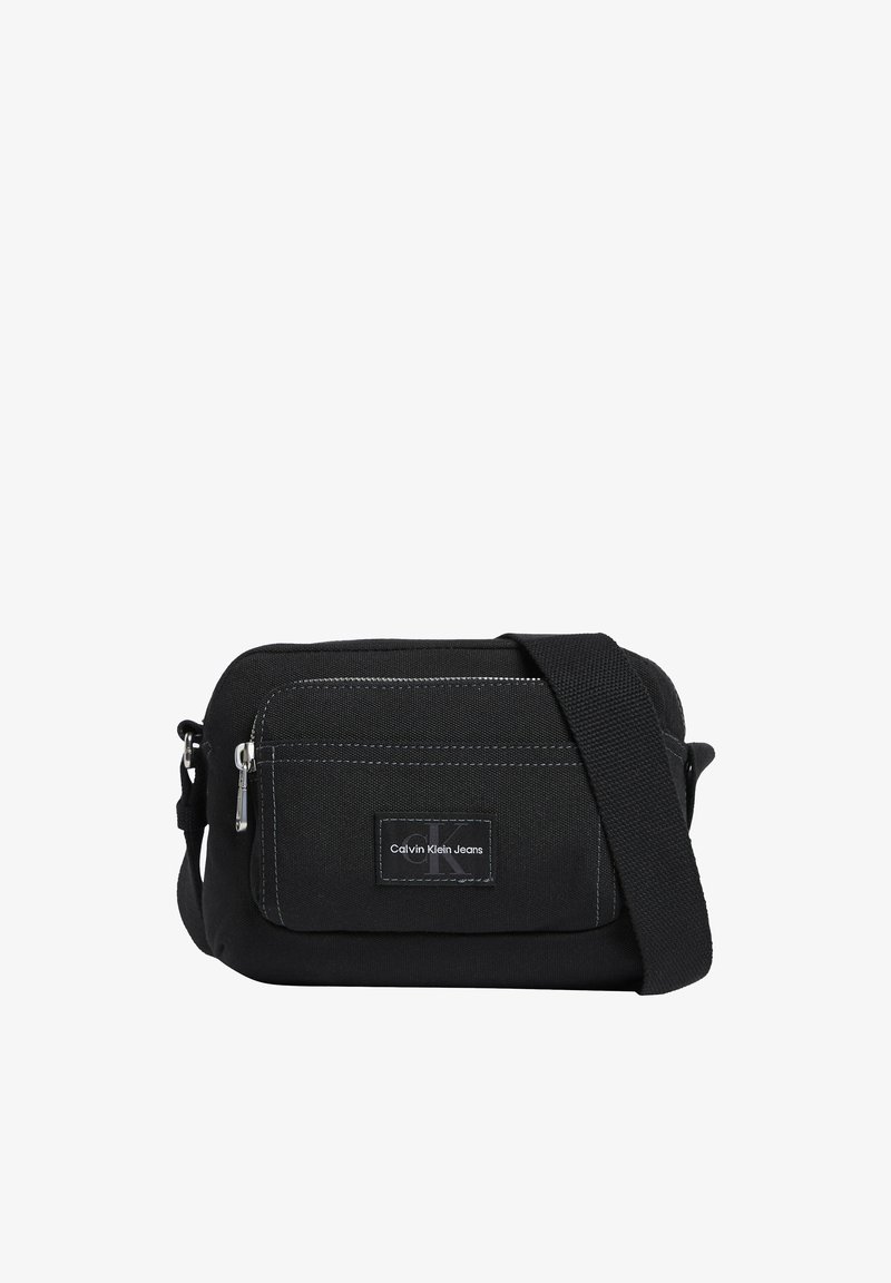 Sac bandoulière en tissu noir avec poche avant zippée, sangle réglable et écusson du logo Calvin Klein Jeans au centre de la poche.