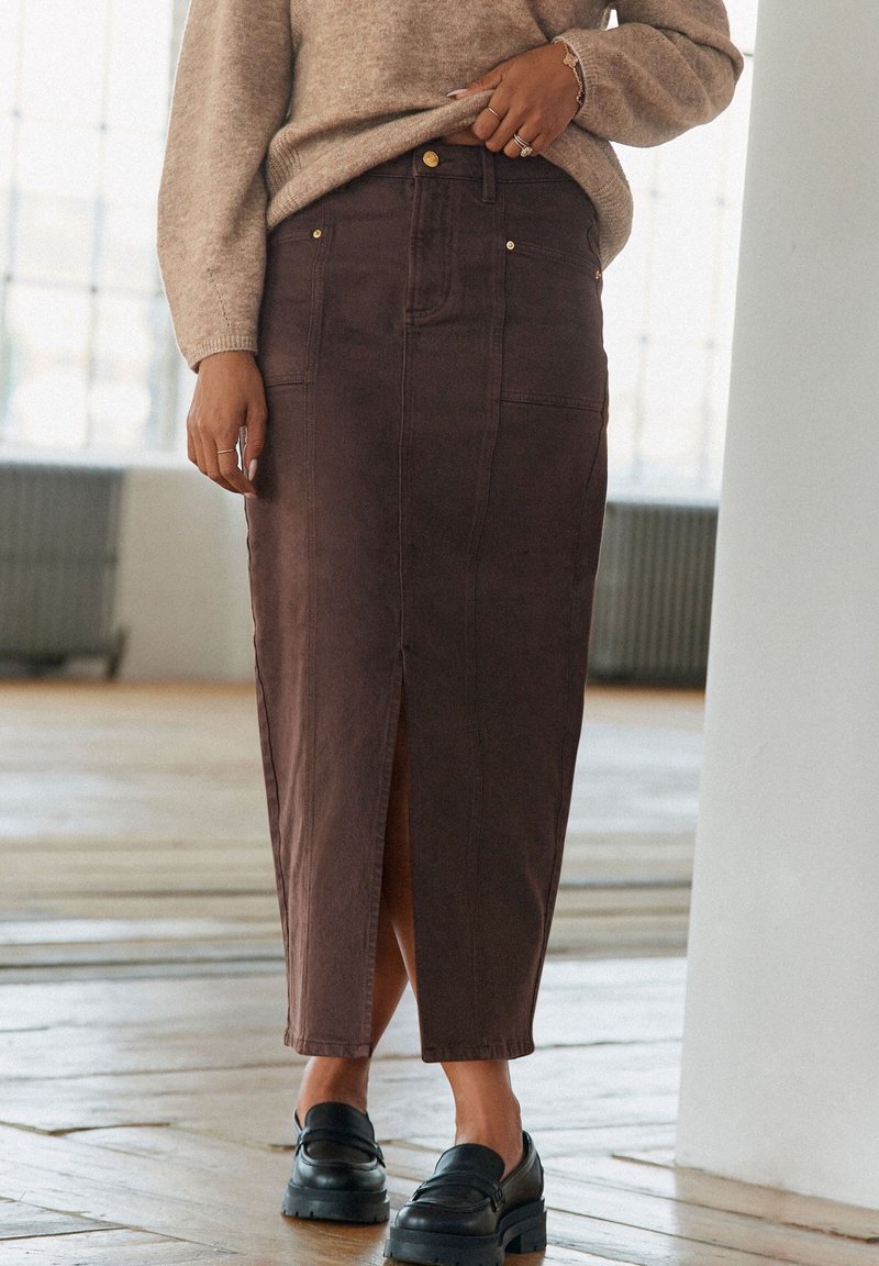 Femme portant une longue jupe marron avec une fente à l'avant, un pull beige, des mocassins noirs et des bagues, debout à l'intérieur sur un sol en bois.