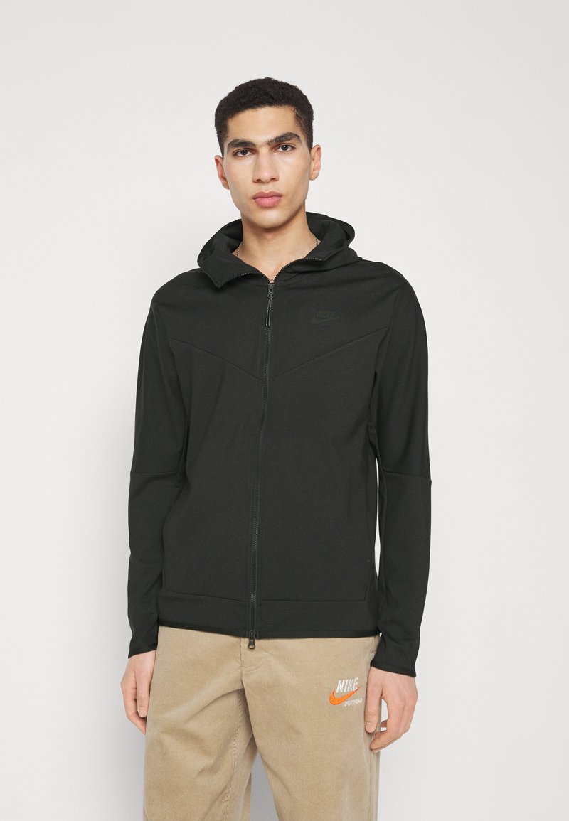Felpa nera con zip e texture liscia, design aderente e cappuccio. Presenta un sottile logo Nike nella parte superiore sinistra e una zip a contrasto.