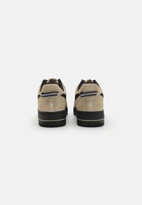 Beige zamšota kedai ar melniem akcentiem, ar mīkstu apkakli un strukturētu gumijas zoli. Aizmugurē ir redzama logotipa birka un pastiprināta šuvuma.