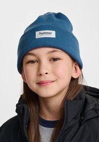 Niebieska dziana czapka z prążkowaną teksturą oraz ma łą białą metką z napisem "hummel sportswear". Noszona przez dziecko, w zestawieniu z czarną kurtką.