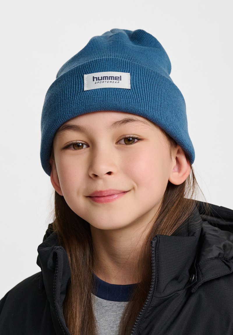 Niebieska dziana czapka z prążkowaną teksturą oraz małą białą metką z napisem "hummel sportswear". Noszona przez dziecko, w zestawieniu z czarną kurtką.