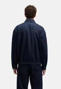 Ciemna kurtka dżinsowa o luźnym kroju, z kołnierzem, elastycznym paskiem w talii i długimi rękawami. Tkanina ma fakturowany, wytrzymały wygląd.