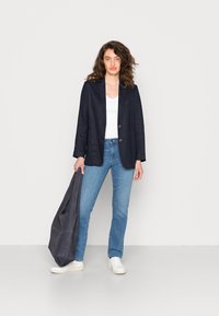 Blazer de linho azul-marinho com dois botões, camisa branca de decote em V, jeans azul claro, bolsa cinza, e ténis brancos, de pé contra um fundo simples.