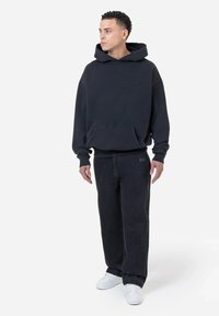 Sudadera negra con capucha y bolsillo frontal, combinada con vaqueros negros de corte suelto y deportivas blancas. Diseño sencillo sin patrones visibles.