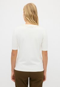 Vit kortärmad topp med ribbad textur, med dekorativa konstpärlor längs halsringningen. Kombinerad med olivgröna byxor.