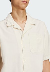 Cream short-sleeve shirt with a textured surface, button-front closure, and a left chest pocket featuring an embossed logo.