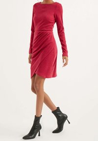 Abito lungo rosso a maniche lunghe con dettaglio drappeggiato, realizzato in un tessuto morbido. Abbinato a stivaletti neri alla caviglia con punta appuntita e tacco alto.