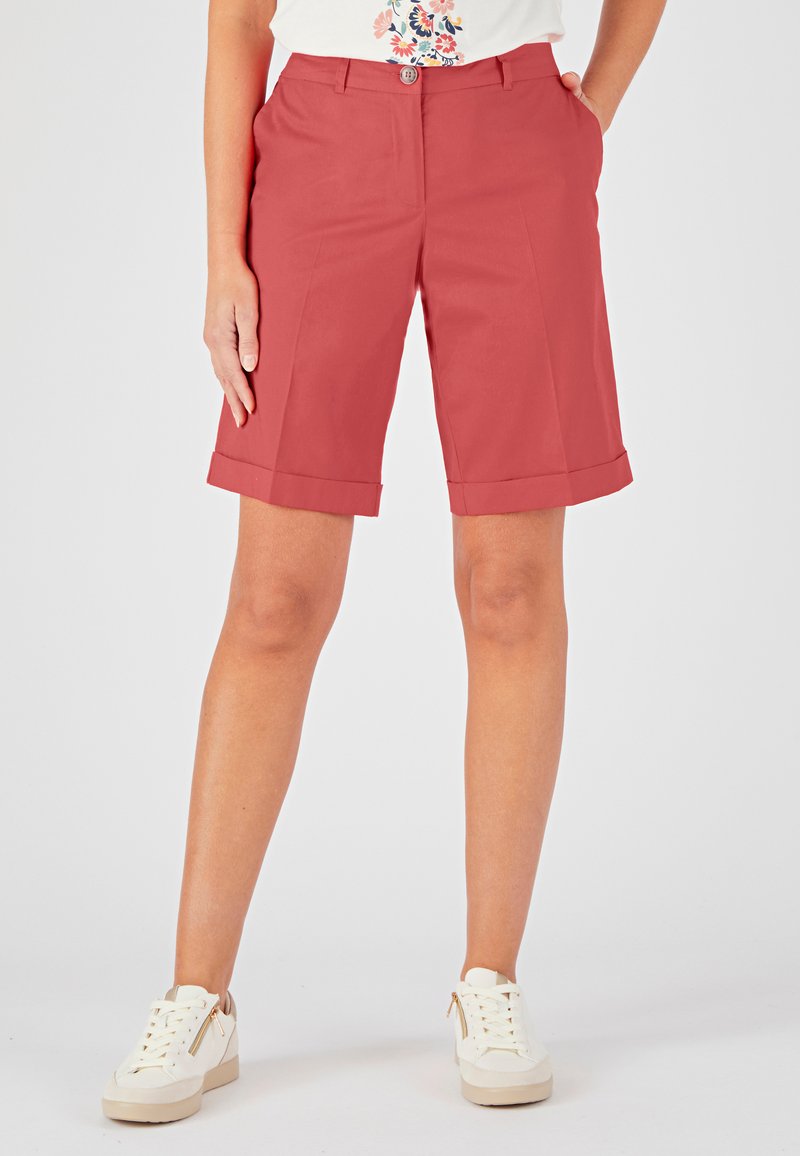 Pantalones cortos de color rosa, ajustados y con dobladillo doblado, cuentan con bolsillos laterales y un cierre de botón, combinados con zapatillas blancas.