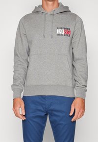 Sudadera gris hecha de una mezcla de algodón con un bolsillo frontal, capucha con cordón y detalles de un logo en rojo, blanco y azul en el pecho.
