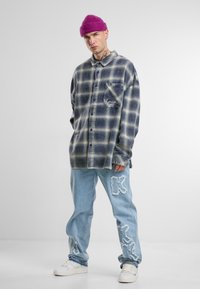 Chemise en flanelle à carreaux bleus et crème avec une poche, associée à un jean bleu clair avec des détails déchirés et un bonnet en tricot rose.