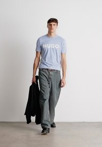 Ανοιχτό μπλε βαμβακερό t-shirt με λευκό λογότυπο "HUGO", συνδυασμένο με γκρι τζιν και ένα μαύρο σακάκι κρεμασμένο στον βραχίονα. Μαύρα παπούτσια.