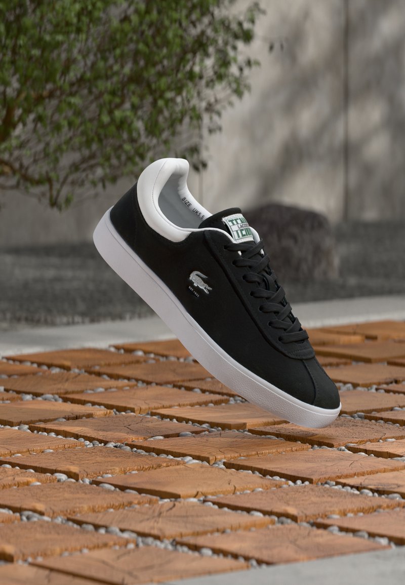 Baskets noires Lacoste avec semelle blanche et logo, flottant au-dessus de carreaux en bois à l'extérieur, plante verte et mur gris en arrière-plan.