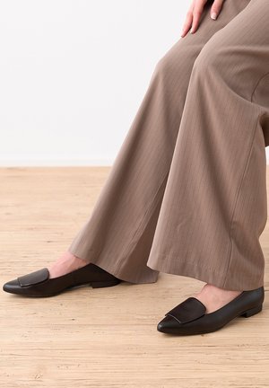 Personne portant un pantalon beige à jambes larges et des chaussures plates en cuir noir sans lacets, assise sur un sol en bois clair devant un fond blanc.