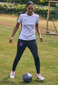 Biele športové tričko Nike s čiernymi akcentmi, v kombinácii s čiernymi nohavicami s ružovým emblémom a bielymi a červenými futbalovými kopačkami, na tráve.