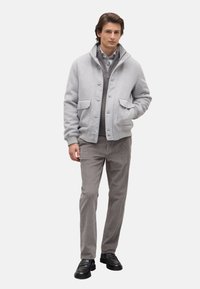 Grijze wolmix jas met een rits- en knoopsluiting, twee voorkockets, gecombineerd met lichtgrijze ribbelbroeken en zwarte schoenen.