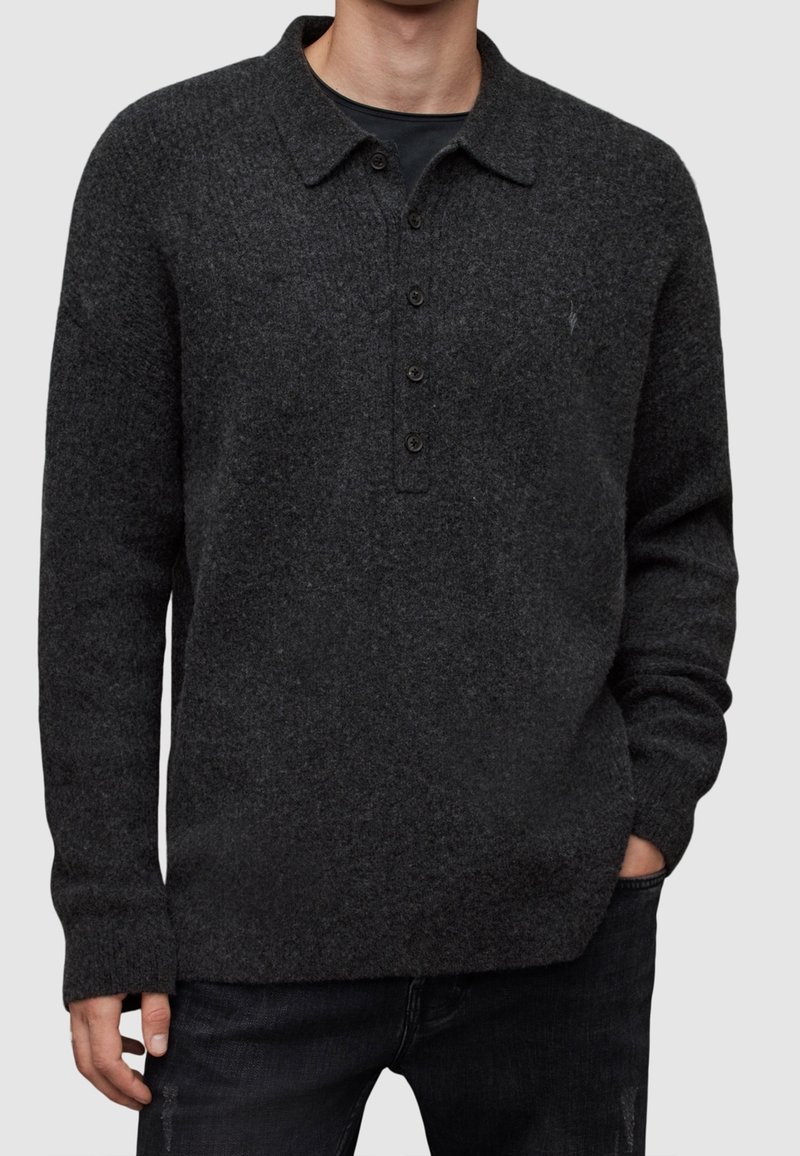 Homme portant un polo en maille gris foncé à manches longues avec quatre boutons et un jean noir, debout avec une main dans la poche.