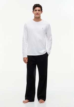 Homme debout pieds nus portant une chemise blanche à manches longues et un pantalon noir ample sur un fond blanc uni.