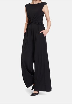 Kvinde iført en ærmeløs sort buksedragt med vide ben, en snoet taljedetalje, hænderne i lommerne og sorte spidse hæle.