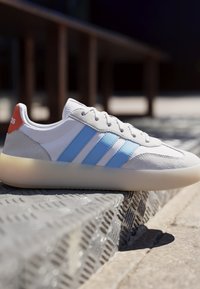 Hvid sneaker med lyseblå striber og en orange hælflap, der hviler på en struktureret metaloverflade i udendørs sollys.