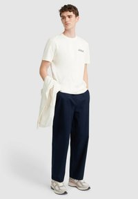 Crème korte mouwen t-shirt met een klein logo, gecombineerd met donkerblauwe broek en lichtgrijze sneakers. Het shirt heeft een gladde textuur.