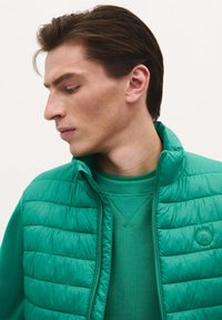 Chaleco acolchado verde con cuello alto sobre una sudadera verde a juego. El material parece suave con una textura acolchada y un detalle de logo en el chaleco.