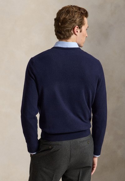 Mand set bagfra iført en navyblå sweater over en lyseblå skjorte og grå bukser mod en ensfarvet beige baggrund.