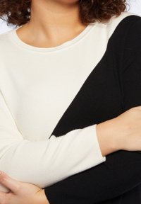 Pull en noir et blanc en blocs de couleur avec une texture côtelée. Il présente un col rond et des manches longues, montrant une position de bras croisée.