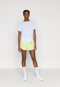 Ανοιχτό μπλε πουκάμισο με κοντά μανίκια και τριγωνικό λογότυπο, συνδυασμένο με σορτς νέον πράσινου χρώματος και λευκά αθλητικά παπούτσια. Η εμφάνιση περιλαμβάνει στοιχεία αθλητικού σχεδιασμού.