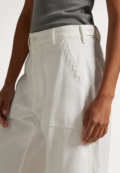 Nærbilde av en person som har på seg høyhøye, hvite bukser med store fremre lommer og en grå, ermeløs, ribbestrikket topp.