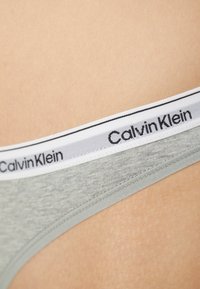 Sivi pamučni tanga s bijelim elastičnim pojasom koji ima crni "Calvin Klein" logo, glatke teksture i minimalističkog dizajna.