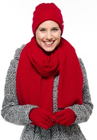 Cappellino lavorato a maglia rosso, sciarpa e guanti abbinati a un cappotto grigio a fantasia. La sciarpa e i guanti sono morbidi, creando un look invernale coerente.