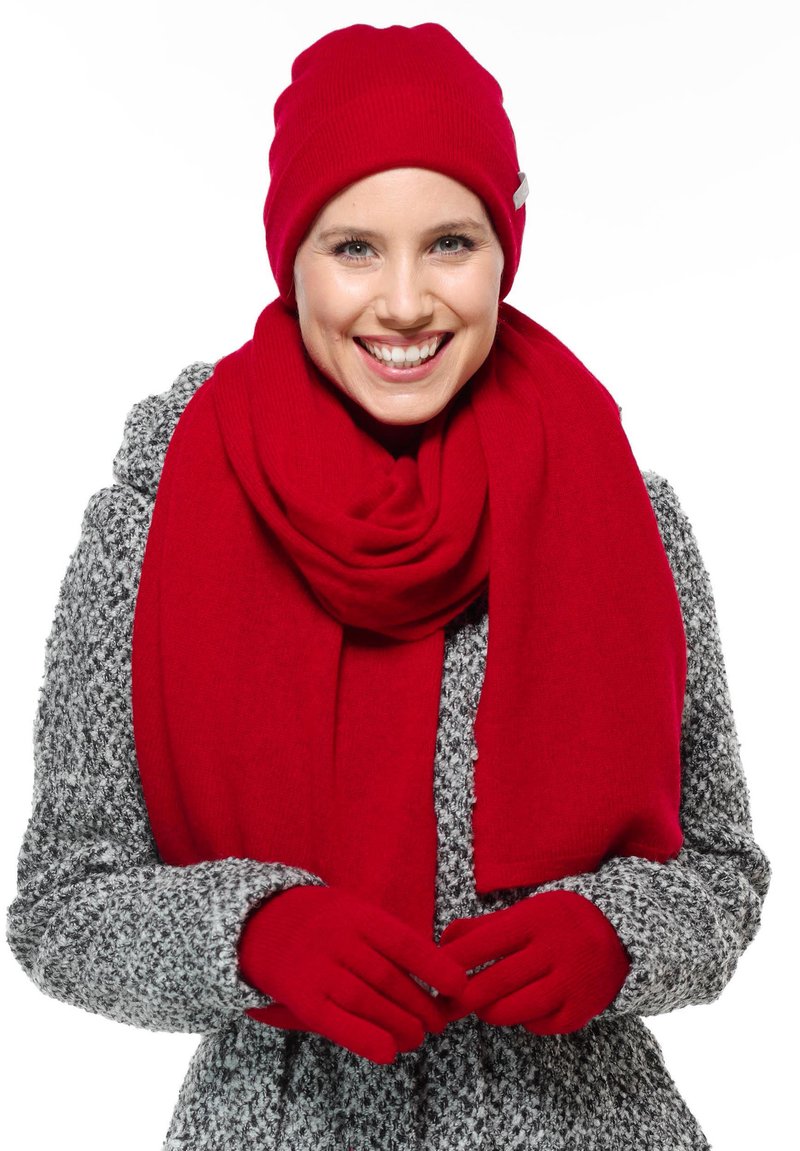 Cappellino lavorato a maglia rosso, sciarpa e guanti abbinati a un cappotto grigio a fantasia. La sciarpa e i guanti sono morbidi, creando un look invernale coerente.