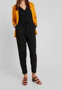 Combinaison noire avec une taille à nouer et un devant boutonné, superposée sous un blazer jaune moutarde vif. Comprend des poches et des pantalons cropped. Talons bruns à lanières.