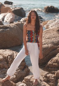 Top senza spalline con un motivo a zigzag in rosso, blu e verde; pantaloni bianchi ampi con cordoncino; modella che si trova su una spiaggia rocciosa.