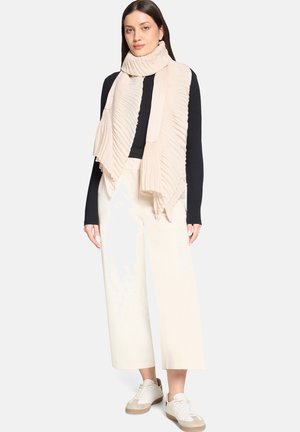 Vrouw met een zwarte top met lange mouwen, een crèmekleurige geplooide sjaal, brede crèmekleurige broek en witte sneakers, staand tegen een effen achtergrond.