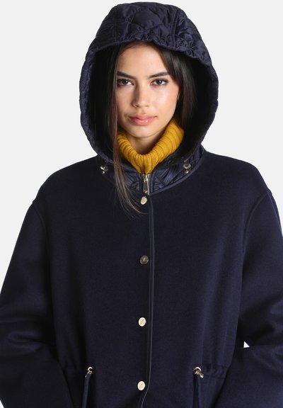 Jeune femme portant un manteau à capuche bleu marine avec des boutons dorés et un pull à col roulé jaune moutarde en dessous, regardant devant elle.