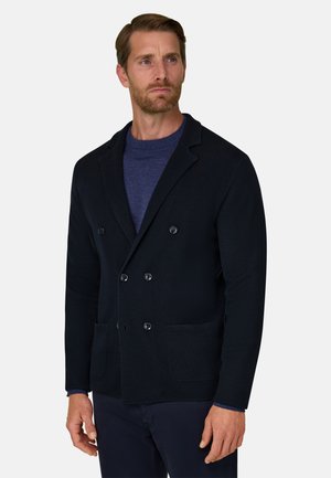 Homme aux cheveux courts et bruns avec une barbe porte un blazer croisé sombre par-dessus un pull bleu, debout devant un fond clair uni.