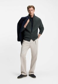 Mörkgrön ribbad zip-up-tröja, svart pikétröja, krämfärgade byxor och svarta sneakers. Man som håller en marinblå kavaj över axeln.