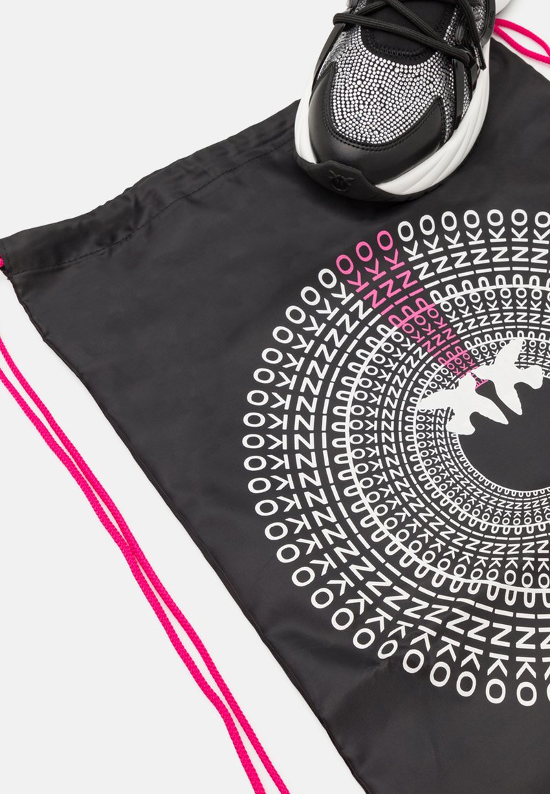 Sac à cordon noir avec un motif graphique circulaire blanc et rose. Comprend des cordons rose vif. Une sneaker noir et blanc est placée sur le dessus.