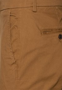 Pantalon en tissu marron à texture lisse, doté d'une poche latérale et d'une fermeture par un seul bouton sur la ceinture.