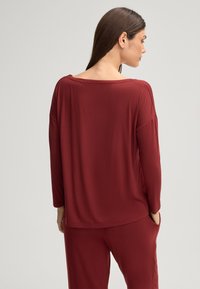 Bordeaux lange mouw top met een wijde halslijn en een relaxte pasvorm. Zachte textuur, heeft verlaagde schouders en zijzakken. Een bijpassende broek is zichtbaar.