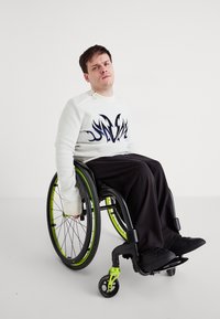 Λευκό πουλόβερ με σχέδιο φλόγας σε μαύρο χρώμα, μαύροι φαρδείς παντελόνες και μαύρα παπούτσια. Το αναπηρικό αμαξίδιο διαθέτει μαύρους τροχούς με πράσινες λεπτομέρειες.