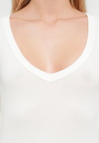 Κοντινό πλάνο ενός ατόμου που φοράει λευκό ριμπ πλεκτό μπλουζάκι με λαιμόκοψη σε σχήμα V, δείχνοντας το ντεκολτέ και το πάνω μέρος του στήθους.